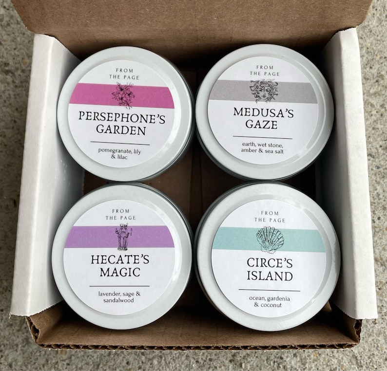 May include: Four small round metal tins with lids, each featuring a different design and scent. The tins are arranged in a 2x2 grid. The top left tin is labeled "Persephone's Garden" with the scent "pomegranate, lily & lilac". The top right tin is labeled "Medusa's Gaze" with the scent "earth, wet stone, amber & sea salt". The bottom left tin is labeled "Hecate's Magic" with the scent "lavender, sage & sandalwood". The bottom right tin is labeled "Circe's Island" with the scent "ocean, gardenia & coconut".