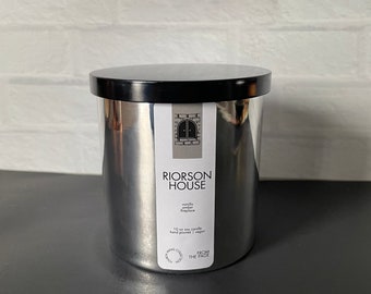 May include: A silver candle jar with a black lid. The jar has a label that says "RIORSON HOUSE" and "Vanilla Ginger Fireplace". The label also says "10 oz soy candle, hand poured, vegan" and "FROM THE FACE".