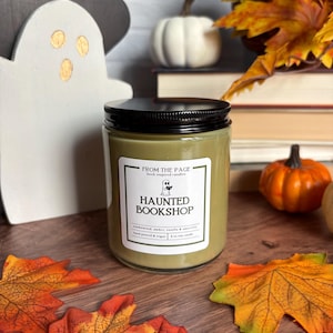 May include: A glass jar candle with a black lid and olive green wax. The white label features the text "FROM THE PAGE book inspired candles" and "HAUNTED BOOKSHOP" with a small ghost illustration, plus "sandalwood, amber, vanilla & amaretto".