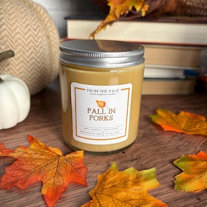May include: A glass candle jar with a silver lid and a tan-colored wax interior. The white label features the text FROM THE PAGE, book inspired candles, FALL IN FORKS, an orange leaf icon, and the text apple, raspberry, cedar & amber, 8 oz soy candle.