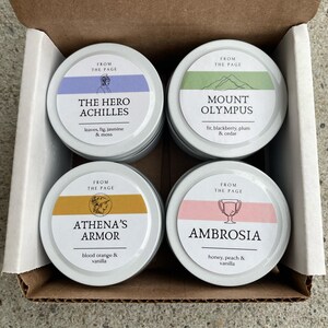 May include: Four small, round, silver candle tins with lids. Each tin has a different colored label with a design and text. The labels read "The Hero Achilles", "Mount Olympus", "Athena's Armor", and "Ambrosia".