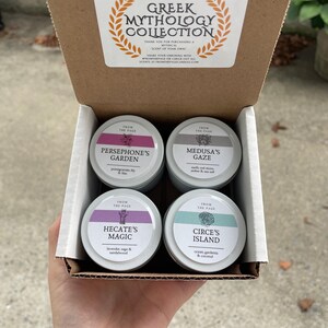 May include: A set of four small, round, metal candle tins with lids. Each tin has a different color lid and features a Greek mythology-themed design and scent description. The tins are arranged in a box with a label that reads "Greek Mythology Collection".