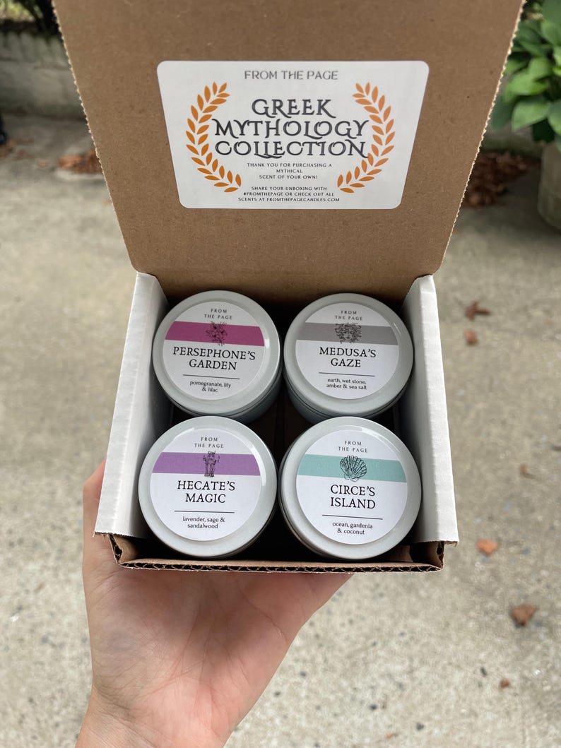 May include: A set of four small, round, metal candle tins with lids. Each tin has a different color lid and features a Greek mythology-themed design and scent description. The tins are arranged in a box with a label that reads "Greek Mythology Collection".