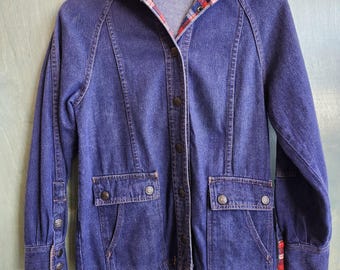 Op de afbeelding: Een vintage spijkerjasje met een met ruitjes gevoerde capuchon en manchetten. De jas heeft knoopsluitingen, twee voorzakken en een taille met trekkoord. De denim is een diep indigokleur, en de ruitjesvoering bevat rood, blauw en wit.