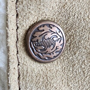 Op de afbeelding: Close-up van een bronzen knoop met een reli&euml;fontwerp van bladeren en een centraal embleem. De knoop is geplaatst tegen een getextureerd, lichtbruin su&egrave;de materiaal, met zichtbare stiksels langs de rand.