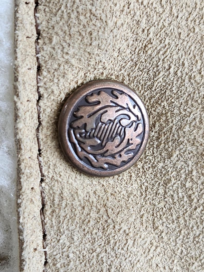 Op de afbeelding: Close-up van een bronzen knoop met een reli&euml;fontwerp van bladeren en een centraal embleem. De knoop is geplaatst tegen een getextureerd, lichtbruin su&egrave;de materiaal, met zichtbare stiksels langs de rand.