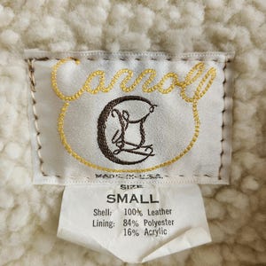 Op de afbeelding: Close-up van een kledinglabel met de merknaam "Carroll" in gouden borduursel. Het label geeft de maat "SMALL" aan en vermeldt de buitenkant als 100% leer en de voering als 84% polyester en 16% acryl. De achtergrond is een cr&egrave;mekleurige, getextureerde stof.