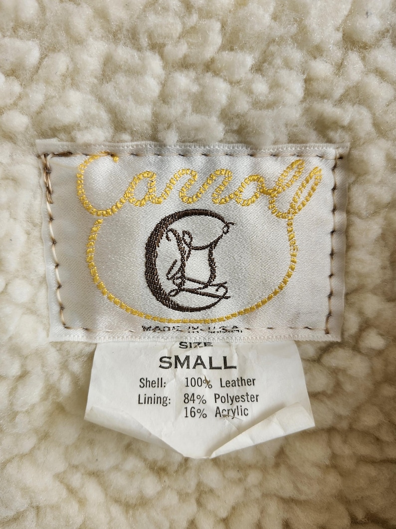 Op de afbeelding: Close-up van een kledinglabel met de merknaam "Carroll" in gouden borduursel. Het label geeft de maat "SMALL" aan en vermeldt de buitenkant als 100% leer en de voering als 84% polyester en 16% acryl. De achtergrond is een cr&egrave;mekleurige, getextureerde stof.