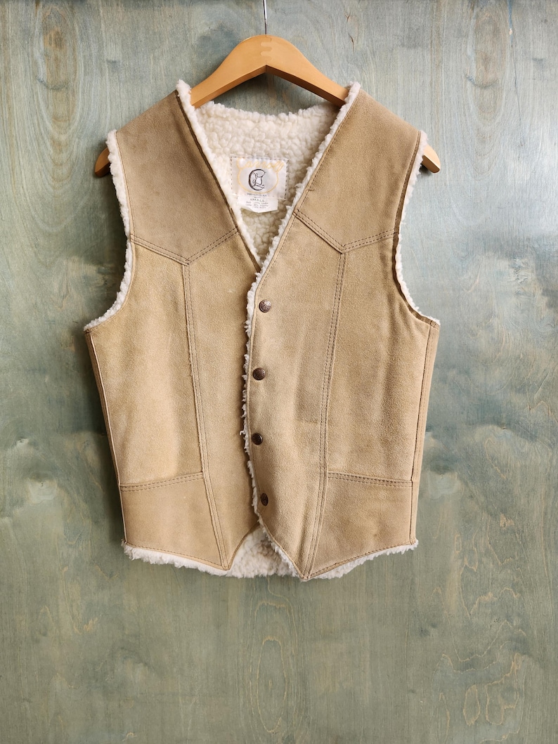Op de afbeelding: Een bruin su&egrave;de vest met een cr&egrave;mekleurige voering en afwerking van schapenvacht. Het vest heeft een V-hals, knoopsluiting en decoratieve stiksels. Het hangt aan een houten kleerhanger tegen een blauwgroene achtergrond.