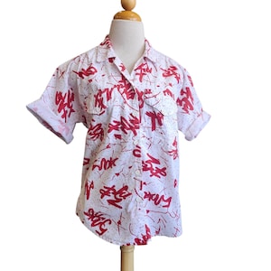 Op de afbeelding: Wit overhemd met rode abstracte print. Het shirt met korte mouwen heeft twee borstzakken en wordt op een mannequin getoond. Het rode ontwerp is een mix van krabbels en vormen.