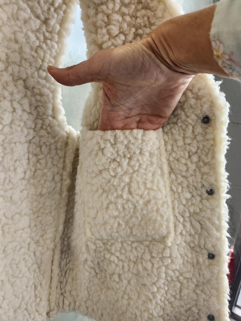 Op de afbeelding: Cr&egrave;mekleurig, pluizig vest met een zak. Het vest heeft een knoopsluiting en een zacht, getextureerd uiterlijk. De zak is zichtbaar en het materiaal lijkt warm en gezellig.