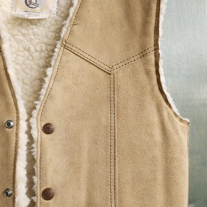 Op de afbeelding: Een bruin su&egrave;de vest met een cr&egrave;mekleurige, donzige binnenvoering. Het vest heeft twee bruine knopen en een decoratief gestikt ontwerp. Een klein label met een logo is aan de binnenkant zichtbaar.