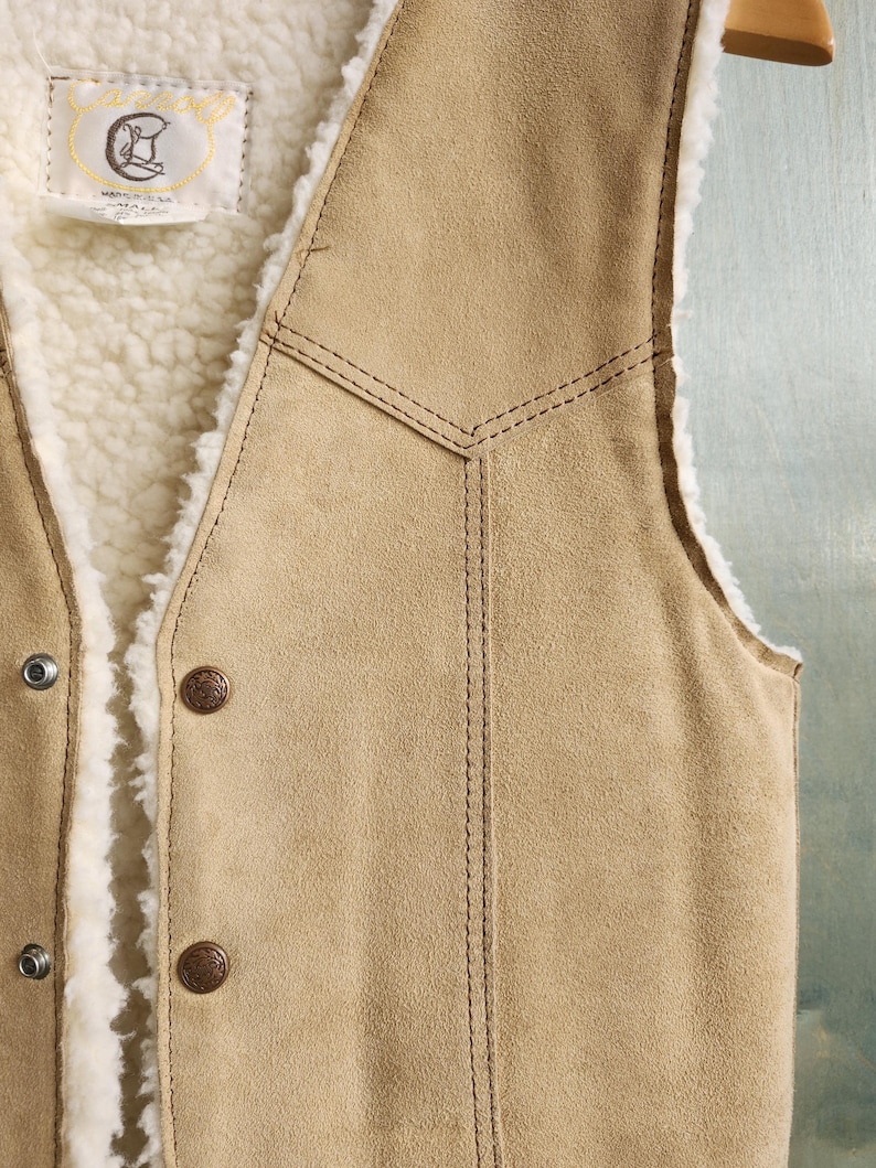Op de afbeelding: Een bruin su&egrave;de vest met een cr&egrave;mekleurige, donzige binnenvoering. Het vest heeft twee bruine knopen en een decoratief gestikt ontwerp. Een klein label met een logo is aan de binnenkant zichtbaar.