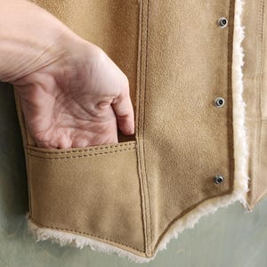 Op de afbeelding: Een bruin su&egrave;de vest met een cr&egrave;me shearling voering. Het vest heeft een zak en metalen drukknoopsluitingen. De stiksels zijn zichtbaar en de stijl is vintage.