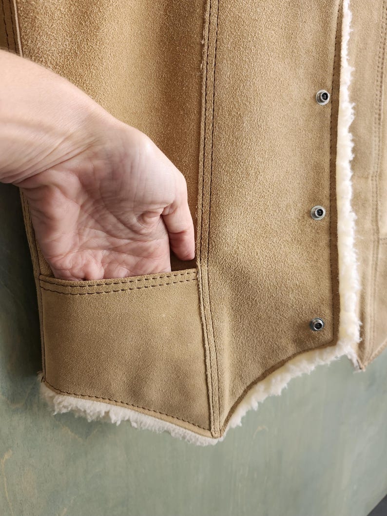 Op de afbeelding: Een bruin su&egrave;de vest met een cr&egrave;me shearling voering. Het vest heeft een zak en metalen drukknoopsluitingen. De stiksels zijn zichtbaar en de stijl is vintage.