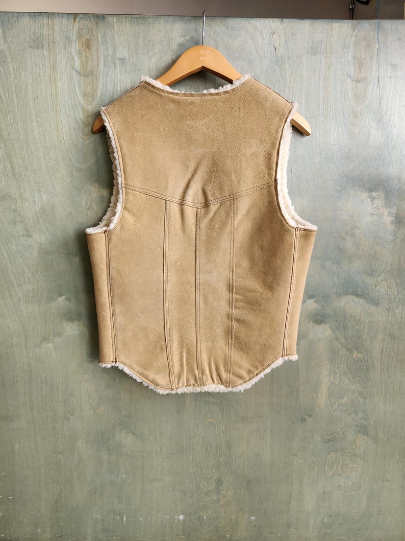 Op de afbeelding: Een bruin vest met een su&egrave;de-achtige buitenkant en een cr&egrave;mekleurige, donzige voering. Het vest hangt aan een houten kleerhanger. De achterkant van het vest heeft verticale stikseldetails en een gebogen zoom.