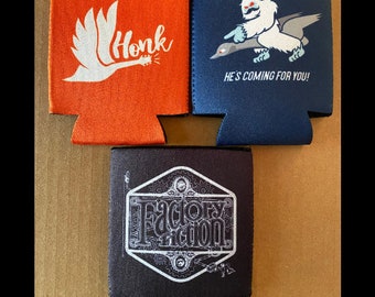 May include: Three different designs of beverage coolers. The first cooler is orange with a white bird graphic and the text "Honk". The second cooler is blue with a white yeti riding a goose and the text "He's coming for you!" The third cooler is black with a white graphic of a gear and the text "Factory Fiction".