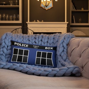 May include: Blue throw pillow with a white "Police Public Call Box" design. The pillow is on a blue knitted throw blanket.