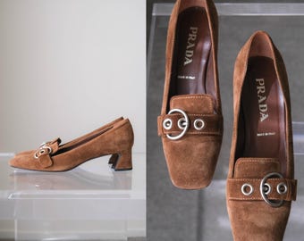 Puede incluir: Un par de mocasines de ante marrón con tacón bajo y punta cuadrada. Cada zapato presenta una tira decorativa sobre el empeine adornada con ojales de metal plateado y una hebilla circular. La plantilla interior lleva la marca Prada.
