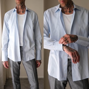 May include: A light blue button-down shirt with long sleeves and a classic collar. The shirt is worn open over a white undershirt and paired with gray trousers. The sleeves are cuffed, revealing the cuff detail.
