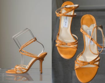 Puede incluir: Un par de sandalias de tacón alto en cuero color amarillo mostaza con tiras finas entrecruzadas adornadas con pequeños remaches metálicos. El calzado incluye una correa ajustable en el tobillo y etiqueta Jimmy Choo.