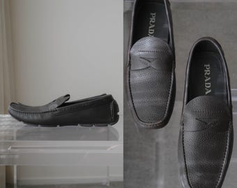 Puede incluir: Un par de mocasines de cuero marrón oscuro con textura granulada. Los zapatos presentan una construcción clásica de tipo mocasín con una tira sobre el empeine, el logotipo de Prada en la plantilla y un detalle de logo sutil en la tira.