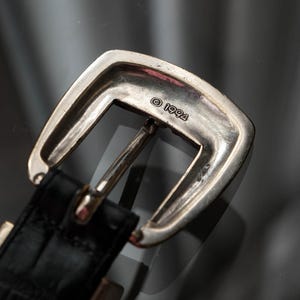 May include: A close-up shot of a silver-coloured belt buckle with a rectangular shape and a curved top. The buckle is attached to a black leather belt with a textured pattern. The year "1994" is stamped on the buckle.