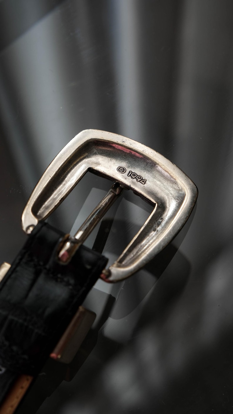 May include: A close-up shot of a silver-coloured belt buckle with a rectangular shape and a curved top. The buckle is attached to a black leather belt with a textured pattern. The year "1994" is stamped on the buckle.