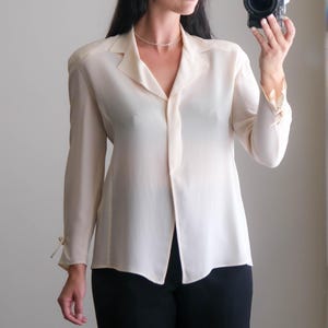 Peut inclure: Un chemisier cr&egrave;me avec un col et des manches longues, associ&eacute; &agrave; un pantalon noir. Le chemisier a un design boutonn&eacute; et des d&eacute;tails de n&oelig;uds aux poignets. La tenue est simple et &eacute;l&eacute;gante.
