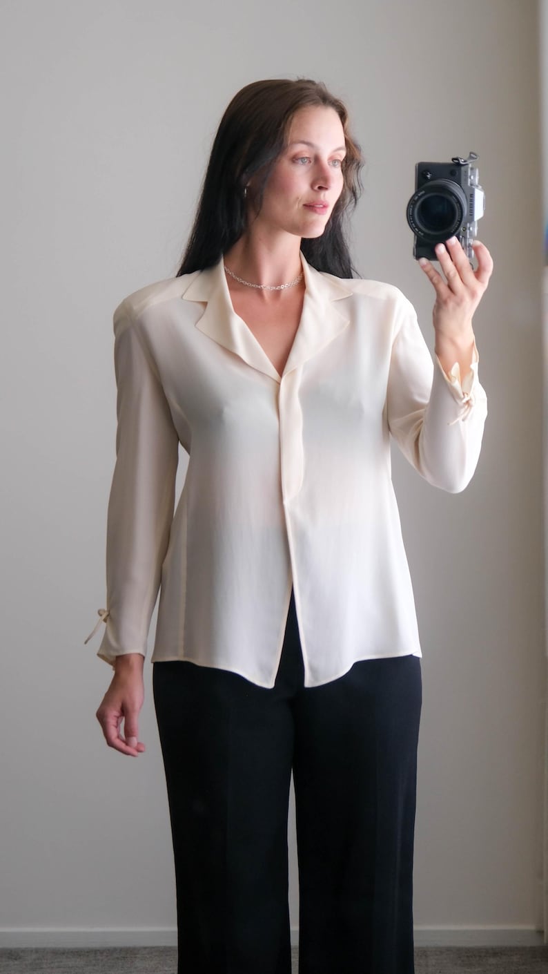 Peut inclure: Un chemisier cr&egrave;me avec un col et des manches longues, associ&eacute; &agrave; un pantalon noir. Le chemisier a un design boutonn&eacute; et des d&eacute;tails de n&oelig;uds aux poignets. La tenue est simple et &eacute;l&eacute;gante.