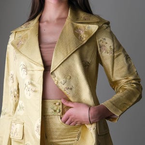 May include: A golden yellow suit with a floral pattern. The suit includes a blazer with a notched collar and two pockets, and matching pants. The fabric has a geometric design. The suit is a vintage style.