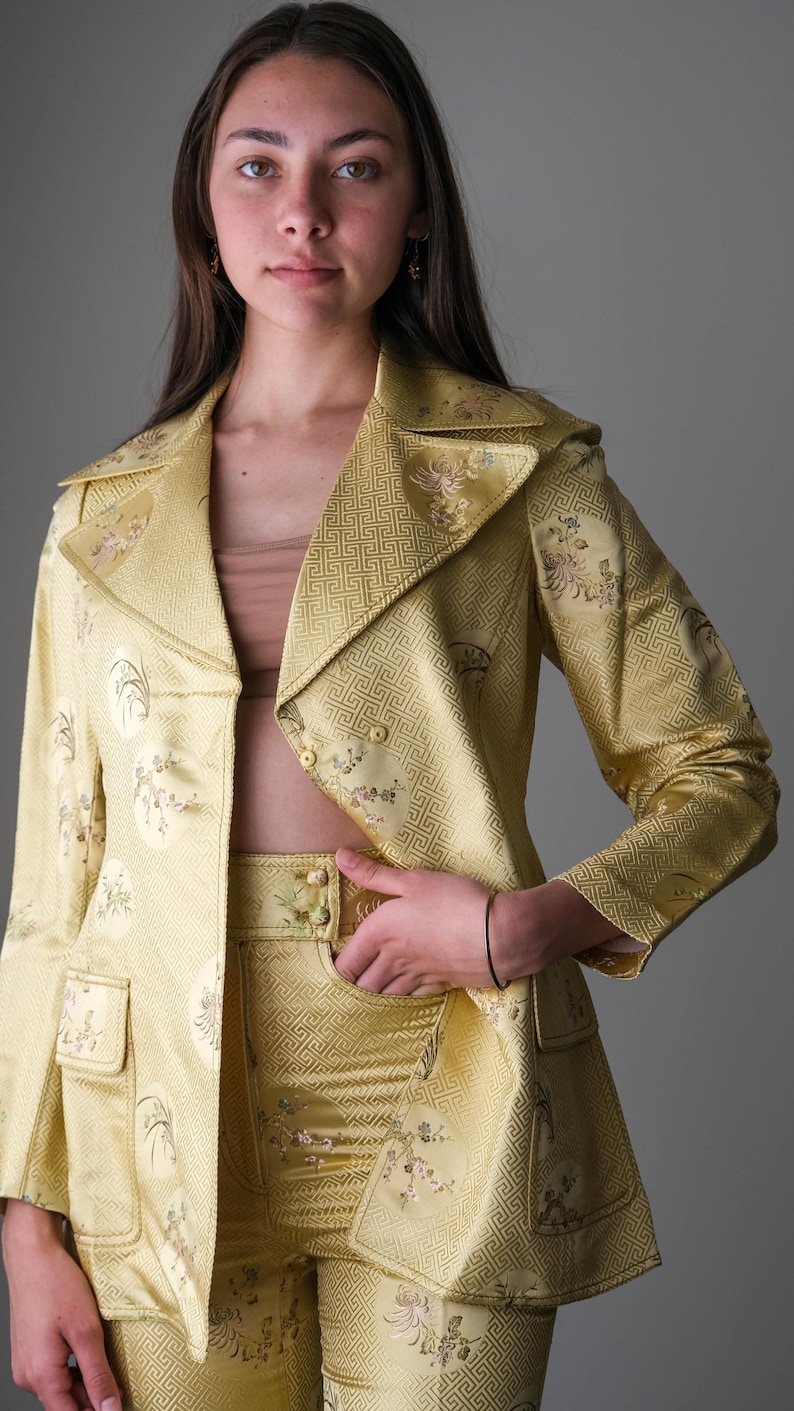 May include: A golden yellow suit with a floral pattern. The suit includes a blazer with a notched collar and two pockets, and matching pants. The fabric has a geometric design. The suit is a vintage style.