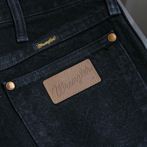Peut inclure: Gros plan sur un jean en denim noir avec une poche arri&egrave;re orn&eacute;e d'un patch "Wrangler" en cuir marron. Le jean a des rivets dor&eacute;s et une petite &eacute;tiquette "Wrangler" dor&eacute;e au-dessus de la poche. Le denim a un aspect l&eacute;g&egrave;rement d&eacute;lav&eacute;.
