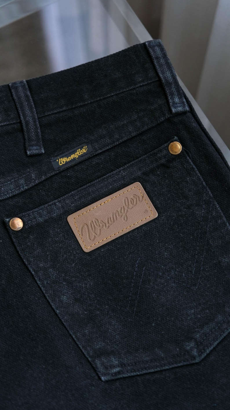 Peut inclure: Gros plan sur un jean en denim noir avec une poche arri&egrave;re orn&eacute;e d'un patch "Wrangler" en cuir marron. Le jean a des rivets dor&eacute;s et une petite &eacute;tiquette "Wrangler" dor&eacute;e au-dessus de la poche. Le denim a un aspect l&eacute;g&egrave;rement d&eacute;lav&eacute;.