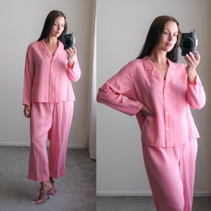 May include: A two-piece pink pajama set. The top is a button-down shirt with a collar and long sleeves. The pants are wide-legged. The set is made of a soft, flowing fabric. The pajamas are a solid, light pink color.