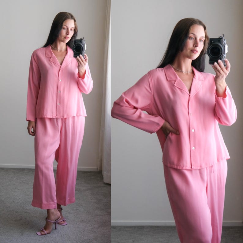 May include: A two-piece pink pajama set. The top is a button-down shirt with a collar and long sleeves. The pants are wide-legged. The set is made of a soft, flowing fabric. The pajamas are a solid, light pink color.