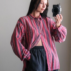 May include: A red, pink, and purple striped button-down shirt with long sleeves and a chest pocket. The shirt is paired with dark blue pants. The model is holding a camera.