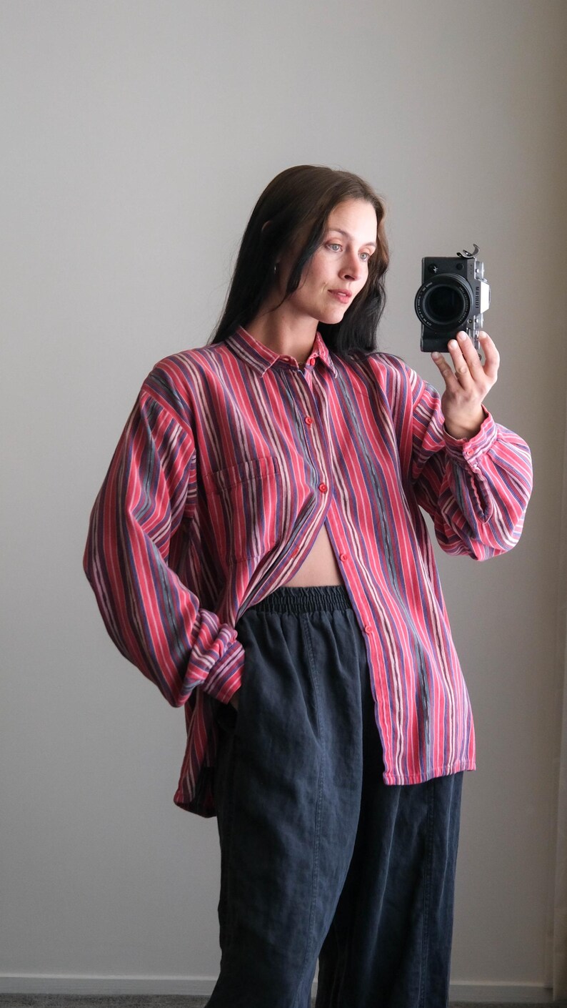 May include: A red, pink, and purple striped button-down shirt with long sleeves and a chest pocket. The shirt is paired with dark blue pants. The model is holding a camera.