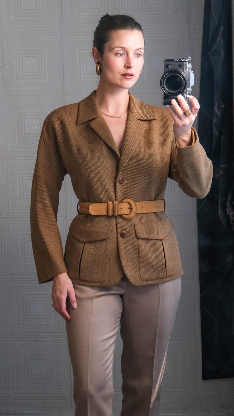 May include: A brown blazer with a matching belt and tan pants. The blazer has two front pockets and a classic collar. The belt cinches the waist. The outfit is a neutral color palette, suitable for a professional or smart casual look.