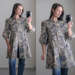 May include: A knee-length coat featuring a round neckline, three large buttons down the front, and three-quarter length puff sleeves. The fabric has a dark gray, tan, and black abstract animal-inspired print with a subtle sheen and a pleated waist.