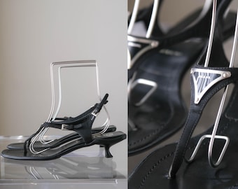 Puede incluir: Sandalias de dedo en cuero negro con tacón bajo tipo kitten y tira trasera con hebilla plateada. La parte delantera de la sandalia muestra una placa metálica triangular con el logotipo de Prada. El diseño incluye una tira vertical fina.
