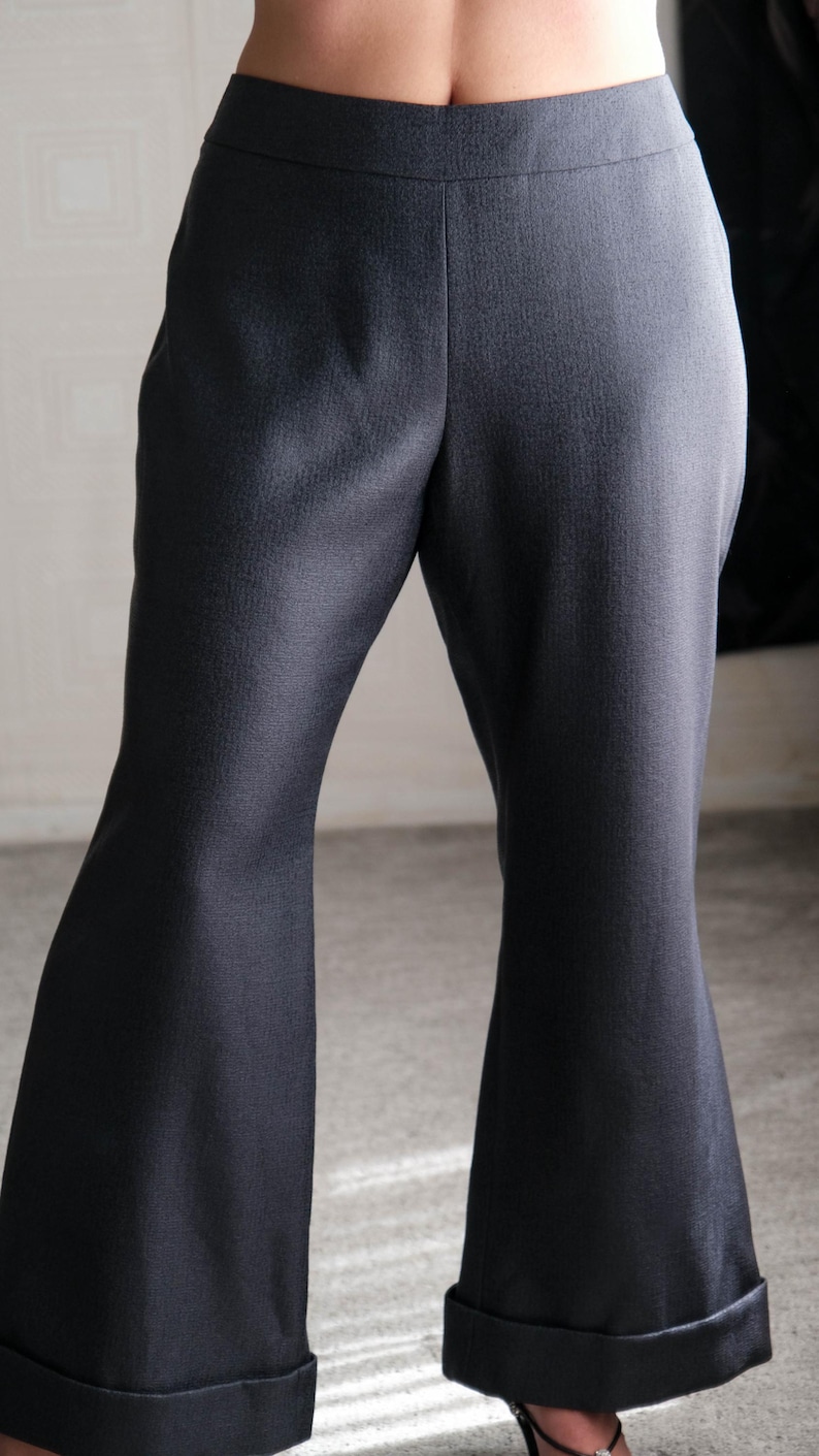 May include: Dark gray flared pants with a tailored fit. The pants have a high waist and a subtle texture. The cuffs are turned up at the hem. The pants are made of a woven fabric.