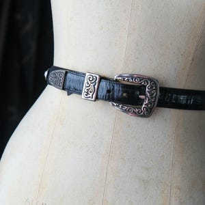 May include: Black leather belt with a silver buckle and decorative accents. The belt is displayed on a mannequin. The buckle features an ornate design. A fashion accessory.