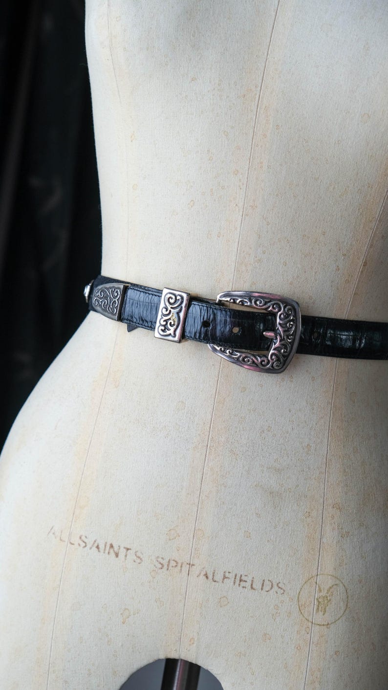 May include: Black leather belt with a silver buckle and decorative accents. The belt is displayed on a mannequin. The buckle features an ornate design. A fashion accessory.