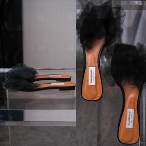 May include: Black slide sandals with a large, ruffled tulle accent. The sandals have a black sole and a tan insole. The brand name "Isaac Mizrahi New York" is visible on the insole. The shoes are displayed on a clear surface.