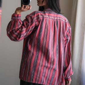 May include: A long-sleeved shirt with vertical stripes in red, purple, and white. The shirt has a button-down front and a classic collar. The sleeves are cuffed at the wrists. The person is wearing dark blue pants.