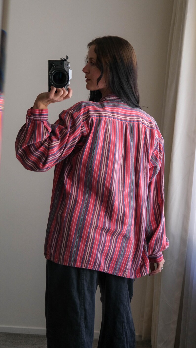 May include: A long-sleeved shirt with vertical stripes in red, purple, and white. The shirt has a button-down front and a classic collar. The sleeves are cuffed at the wrists. The person is wearing dark blue pants.