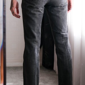 May include: A pair of black denim jeans with a straight leg fit. The jeans have a classic five-pocket design and a leather patch on the back waistband with the word "Wrangler" printed on it.