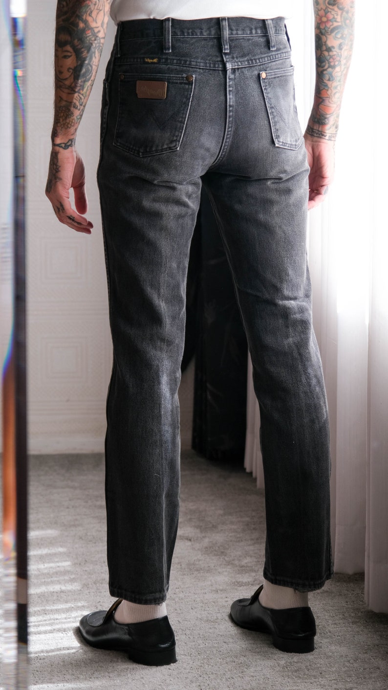 May include: A pair of black denim jeans with a straight leg fit. The jeans have a classic five-pocket design and a leather patch on the back waistband with the word "Wrangler" printed on it.