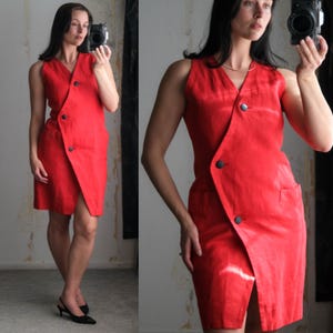 May include: A sleeveless, knee-length red dress with a wrap-style front and black buttons. The dress features a V-neck and a slit in the front, with a pocket on the side. The fabric appears to be a lightweight material.